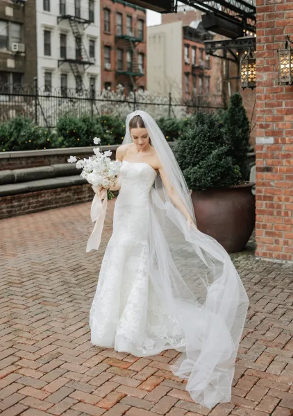 Bridal portrait of a bride in a strapless lace wedding dress holding a white bouquet with ribbon, looking down in a brick cobblestone courtyard