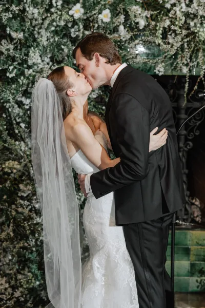 Wedding kiss portrait of bride and groom kissing, her long veil and lace strapless gown framed by a white flower and greenery wall