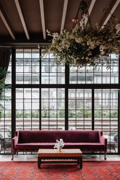 Wedding lounge decor with a burgundy velvet sofa, coffee table, and candles under a hanging floral chandelier by black grid windows in a courtyard venue
