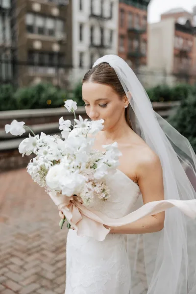 Bridal portrait of a bride holding bouquet as she smells white flowers in a strapless lace dress and cathedral veil on a brick city walkway