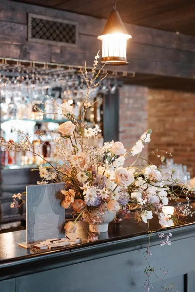 Bar wedding florals with a wedding bar decor centerpiece of pastel roses and wildflower stems in a ceramic vase on a wood bar, lantern glow against brick shelves