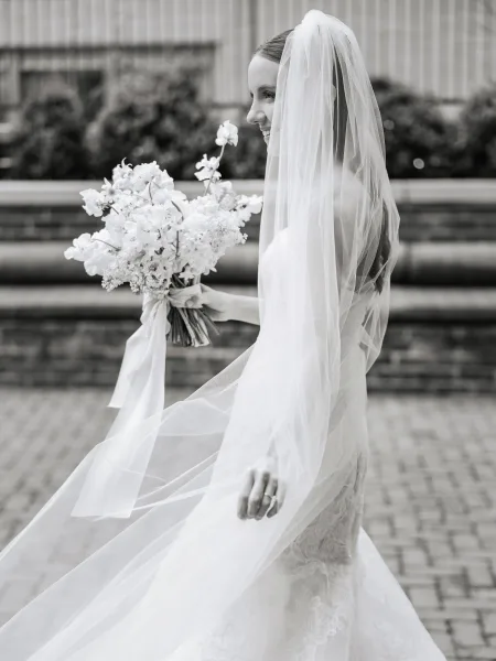 Bridal portrait in black and white of a smiling bride in side profile holding a ribbon-wrapped bouquet, veil blowing by a brick wall