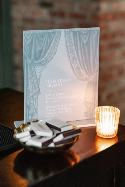Signature drinks sign on a table with a wedding signature drinks menu, acrylic display, candle votive, matchbooks, and brick wall backdrop