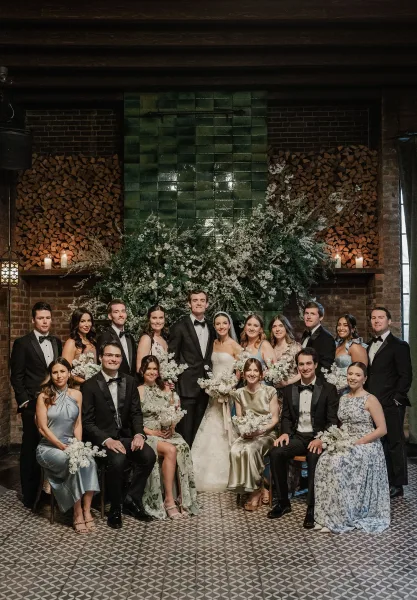 Wedding party portrait with bride and groom seated by a brick fireplace, bride holding white bouquet amid greenery and candle décor indoors