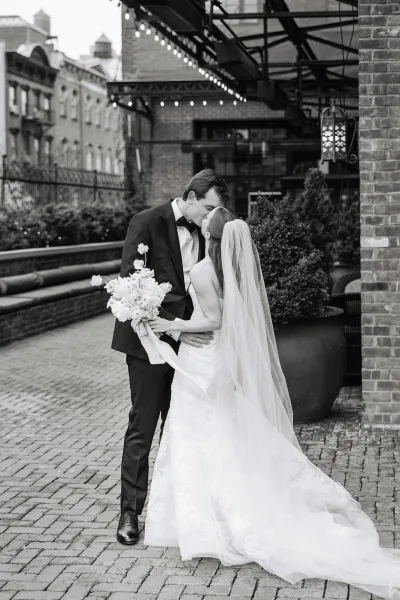 Wedding kiss portrait of bride and groom kissing, her long veil and bouquet against brick courtyard with cobblestones and string lights