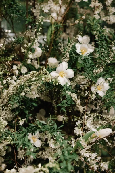 Wedding florals of white wedding flowers with flowering branches and lush greenery against dark foliage and blurred reception seating