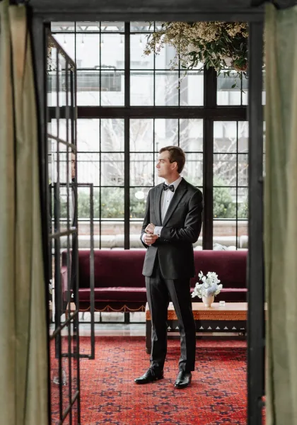 Groom portrait in a black tuxedo with bow tie and boutonniere, seated by large grid windows in an indoor lounge with velvet sofa