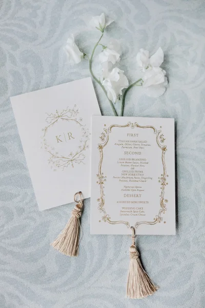 Wedding menu card with gold foil lettering and ornate border, finished with a tassel, styled on light blue patterned fabric