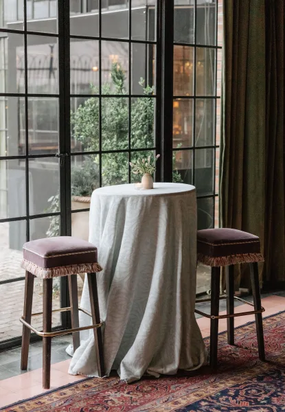 Cocktail table setup with white tablecloth, small floral centerpiece and votive candle, flanked by bar stools near grid windows and greenery