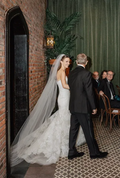 Wedding processional as bride walking down aisle in a lace gown and cathedral veil, escorted past seated guests in a brick-walled room