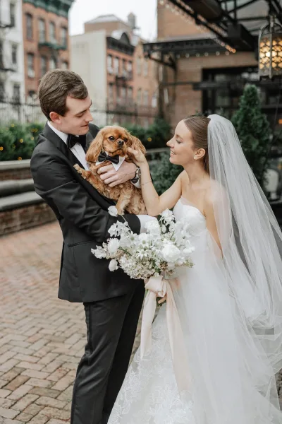 Couple portrait with dog as bride in veil and groom in tuxedo hold a bow-tied pup in a brick courtyard with string lights