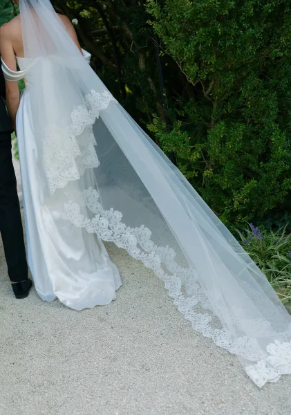 Bridal veil, a delicate lace edge veil, flows behind an off-the-shoulder gown as the couple walks a gravel garden path amid lush greenery.