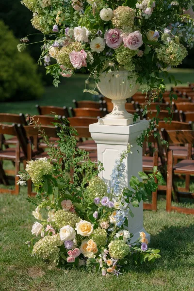 Ceremony flowers in an urn vase define the garden ceremony decor, a pastel arrangement on a grass lawn framed by leafy garden trees.