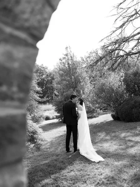 Wedding kiss portrait in black-and-white, the bride with a long veil kisses her groom beneath garden trees beside a low stone wall.