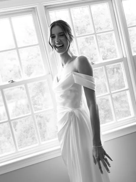 Bridal portrait of a bride by the window, laughing with an off-the-shoulder neckline, framed by large white windows and garden trees.