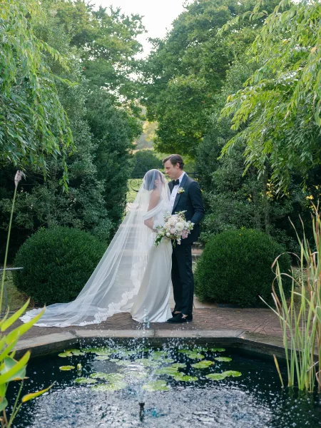 Couple portrait of a bride and groom in garden with her long veil trailing as they stand by a fountain and lily pond along a brick path.