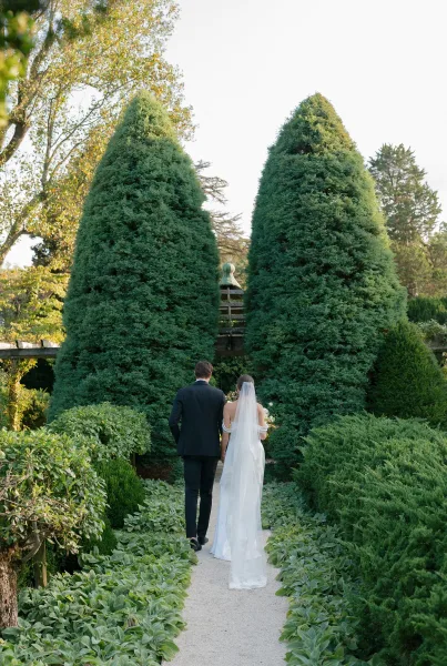 Couple portrait of the bride and groom walking along a gravel garden path edged by topiary, her long veil trailing behind them.