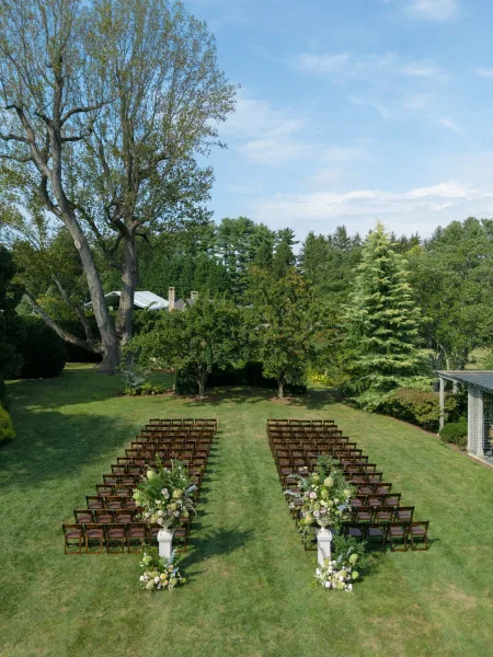 Outdoor ceremony setup with garden ceremony seating in neat rows of wooden chairs on a lawn, facing a pergola amid garden trees and blue sky.