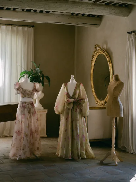 Bridal gown display with wedding dress on mannequin beside floral print dresses, framed by an ornate gold mirror in rustic window light