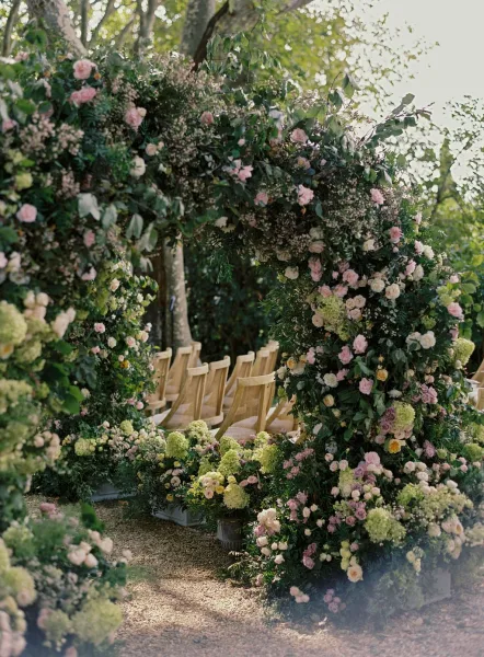 Ceremony aisle decor with a floral ceremony arch of roses, hydrangeas, and greenery garland lining a sunlit gravel garden path with wooden chairs
