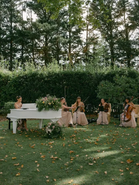 Wedding musicians perform live beside a white grand piano with string instruments in blush dresses on a garden lawn under tall trees