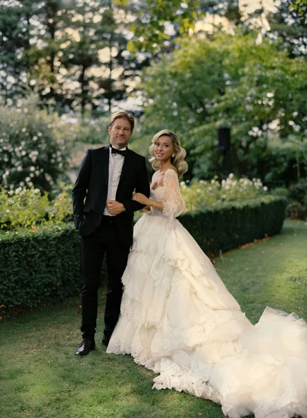 Couple portrait of bride holding the groom’s arm, her lace wedding dress with long train flowing as they stand on a garden lawn