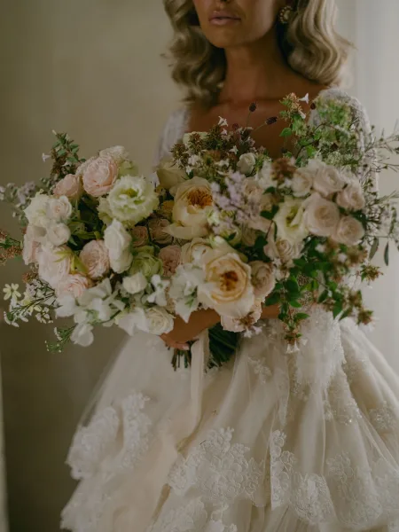 Bridal portrait of a bride holding bouquet, showing ivory lace long-sleeve wedding dress and ribbon-wrapped blooms against a neutral wall