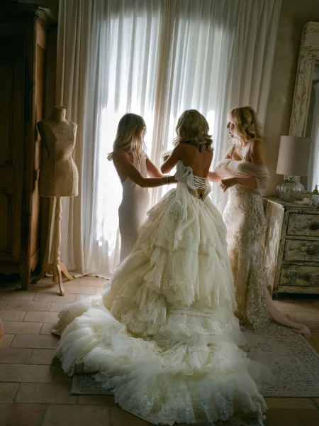 Bridal getting ready as bridesmaids lace a corset back wedding dress with a long tulle train in a window-lit rustic bedroom