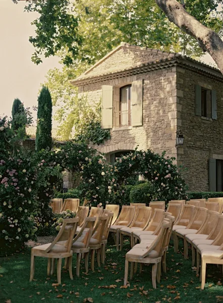 Ceremony setup with outdoor ceremony seating of wood chairs facing a rose and greenery floral arch on a garden lawn by a stone house