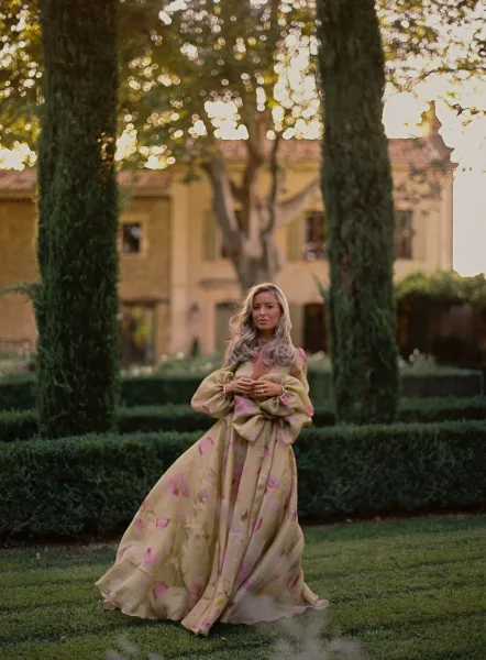 Bridal portrait of a bride in a floral print, off-the-shoulder gown with puff sleeves, holding her skirt in a sunset villa garden with cypress trees