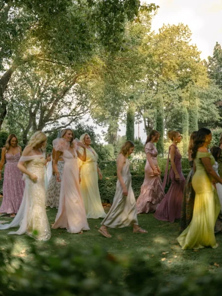 Bridesmaid procession in pastel dresses walking down the aisle, sunlight on a garden lawn with trees, one holding a bridal veil