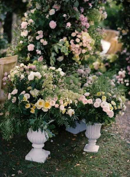 Wedding floral arrangement of pastel roses, greenery, and hydrangea in white urns lining a garden aisle with wooden chairs on grass