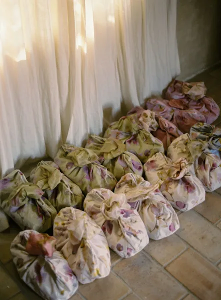 Wedding favor bags wrapped in floral print cloth, tied in neat knots, arranged on a tiled floor by sheer curtains and window light