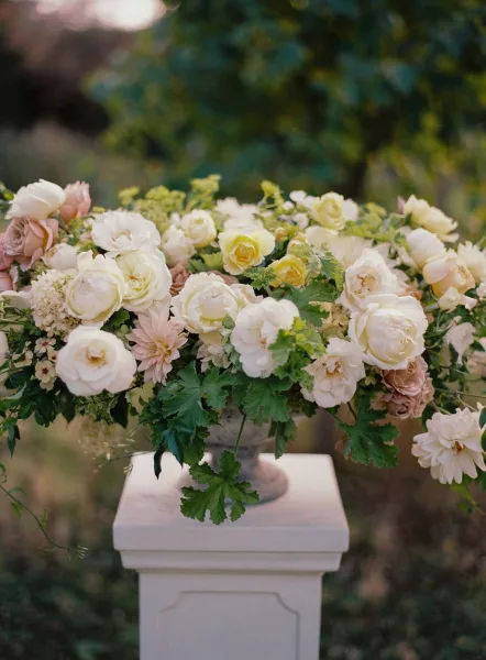 Wedding floral arrangement in a stone urn filled with blush garden roses, ranunculus, and greenery on a white pedestal in sunlight