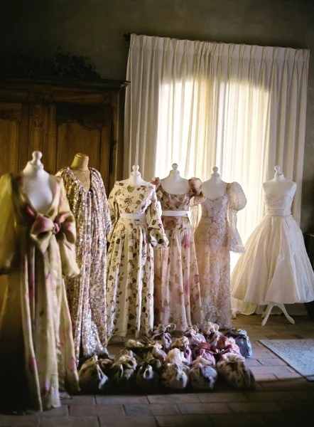 Wedding dress display with bridal gown and floral, sequin, and lace dresses on mannequins by a curtained window and wooden armoire