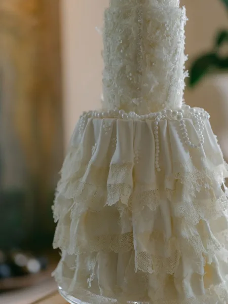 Wedding cake with lace wedding cake ruffles, pearl garland, and textured ivory tiers on a table with greenery against a neutral wall