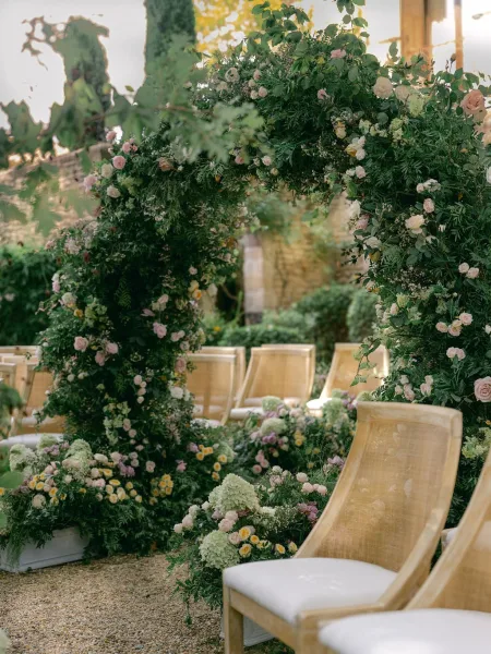 Wedding ceremony arch adorned with roses and greenery garland, grounded florals lining the aisle in a sunlit garden setting