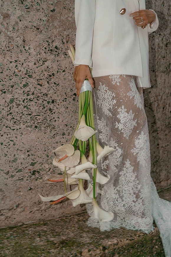 Bridal bouquet with white calla lily bouquet and anthurium blooms, ribbon-wrapped stems held against lace dress by stone wall