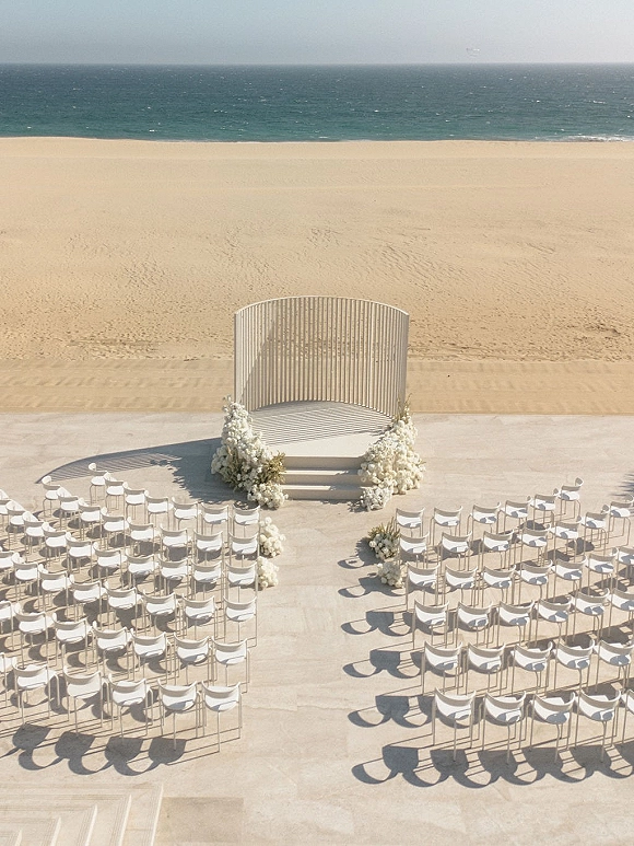 Beach ceremony setup with white wedding altar and round slat backdrop, aisle florals, and white chairs facing the ocean horizon