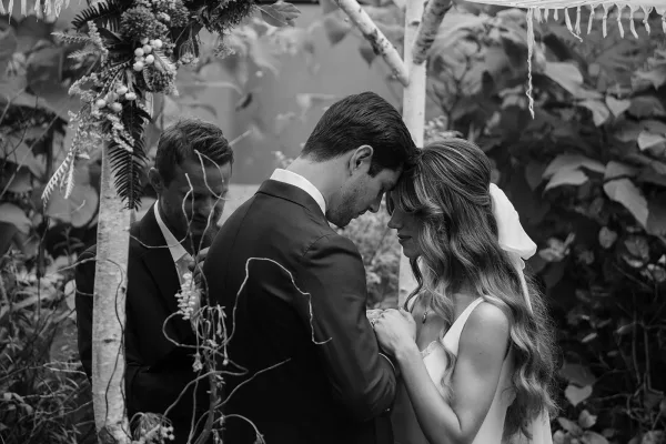 Wedding vows as bride and groom touch foreheads and hold hands, simple veil accent, officiant behind under greenery and ribbons