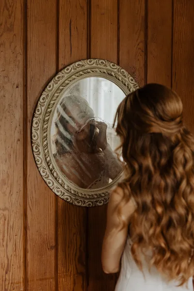 Bridal portrait of a bride in mirror reflection, wearing a wedding dress with braided half-up hair by a wood-paneled wall in window light