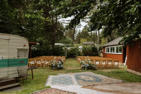 Ceremony setup with outdoor ceremony seating on wood folding chairs facing a layered rug aisle with florals and bunting in forest clearing