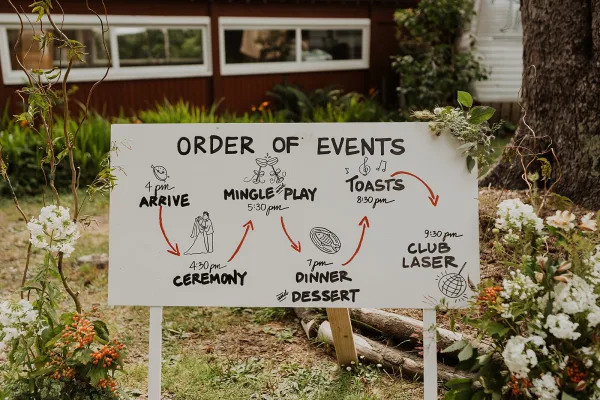 Wedding schedule sign with hand-lettered order of events on an easel, framed by greenery, white flowers, and orange berries on a lawn