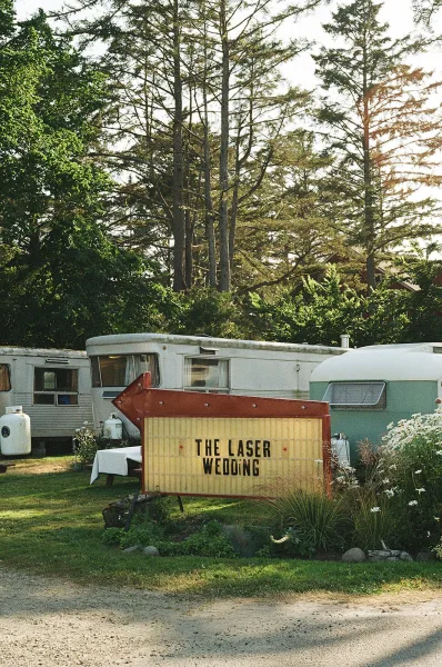 Wedding signage with marquee wedding sign and arrow letters on a white-draped table, wildflowers nearby, set by travel trailers in sunlight