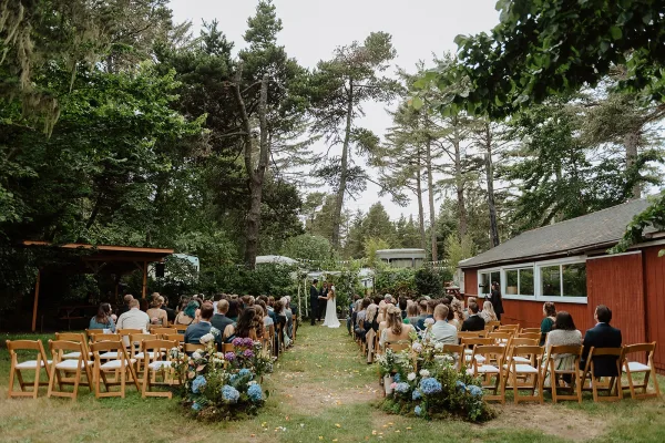Outdoor wedding ceremony with wood folding chairs lining a grass aisle, hydrangea floral arrangements, and bunting beneath tall pines