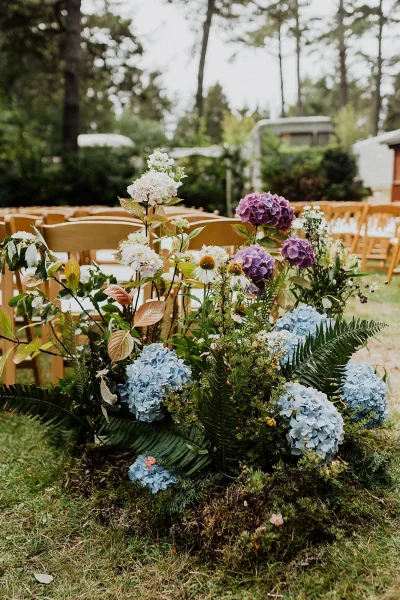 Ceremony aisle florals with blue and purple hydrangeas and fern greenery lining wooden folding chairs on a grass lawn under trees