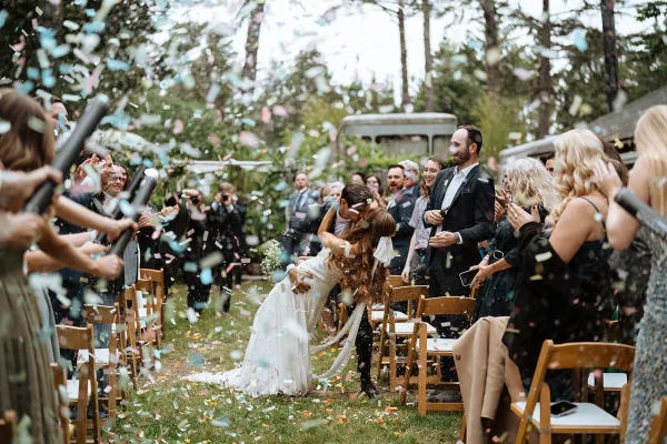 Wedding recessional with confetti wedding exit as bride and groom kiss on the aisle, guests cheering in a garden with trees and a vintage bus behind
