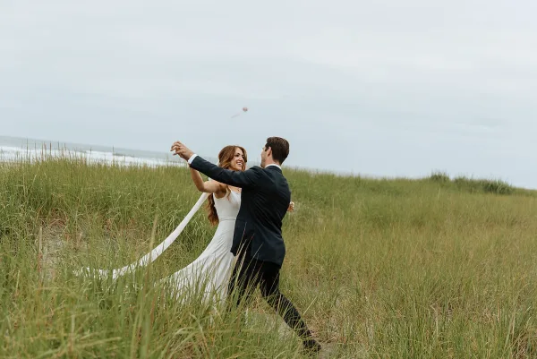 Couple portrait of bride and groom twirling, her long veil trailing over sand dunes with beach grass and a misty ocean backdrop