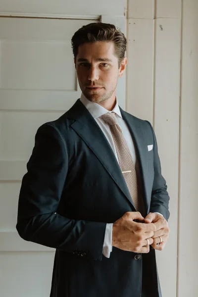 Groom portrait in a dark suit with textured tie, tie bar, and pocket square, showing wedding band against a white paneled wall indoors