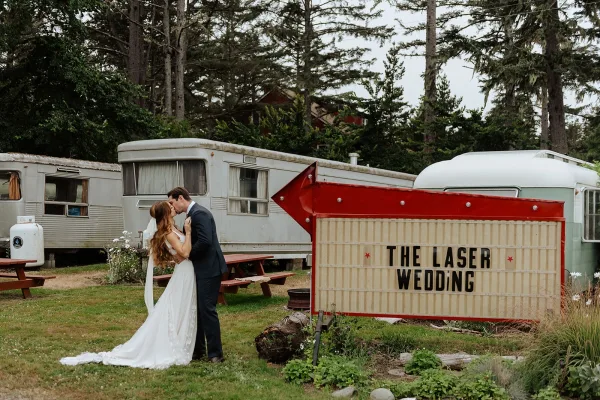 Wedding kiss as bride and groom embrace, her long veil flowing beside a marquee sign on a grassy lawn with vintage trailers